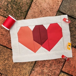 May include: A white fabric panel with a patchwork heart design. The heart is made of red, pink, and burgundy fabric pieces. The panel is pinned with colourful clips.
