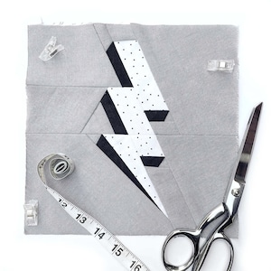May include: A gray fabric square with a black and white lightning bolt design. The fabric is pinned with clear plastic clips. A measuring tape and a pair of silver scissors are also in the image.