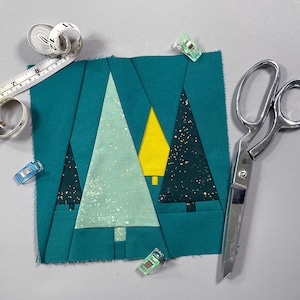 Könnte beinhalten: Ein Patchwork aus türkisfarbenem Stoff mit drei unterschiedlichfarbigen Dreiecken, die ein Weihnachtsbaumdesign bilden. Der Stoff ist zugeschnitten und bereit zum Nähen. Auf dem Bild sind auch Nähklammern und eine Schere zu sehen.