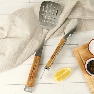 May include: A stainless steel slotted spatula and a pair of tongs with light wood handles. Both tools feature the engraved text "CHIEF OF THE CHARCOAL Happy Father's Day - Love Charlie" and the brand name "ECOLOGY" printed on the metal shafts.