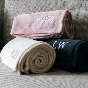 May include: Three rolled up plush blankets with embroidered initials. The blankets are in shades of pink, beige, and black.