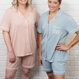 May include: Matching pajama sets in light pink and blue-green. Each set includes a short-sleeved button-down shirt and matching shorts, trimmed with white piping. The shirts are monogrammed with white initials. The pajamas are made of a soft, comfortable material.