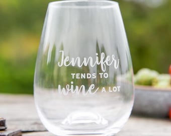 May include: A clear stemless wine glass with the text "Jennifer tends to wine a lot" in white script. The glass is on a wooden surface with a blurred green background.