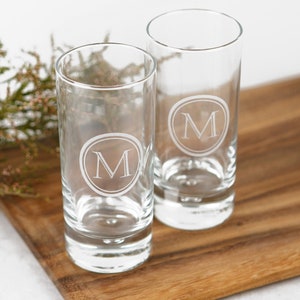 May include: Two clear glass shot glasses with a round, etched design featuring the letter "M" in the centre. The glasses are on a wooden surface.