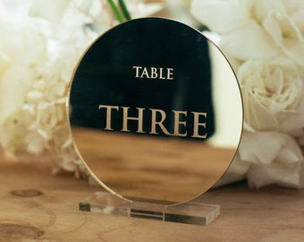 May include: A gold round mirror table number with the text "TABLE THREE" on a clear acrylic stand. The table number is surrounded by white flowers.