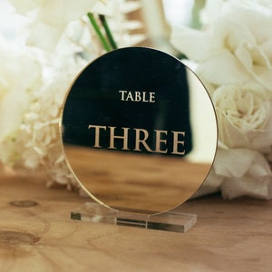 May include: A gold round mirror table number with the text "TABLE THREE" on a clear acrylic stand. The table number is surrounded by white flowers.