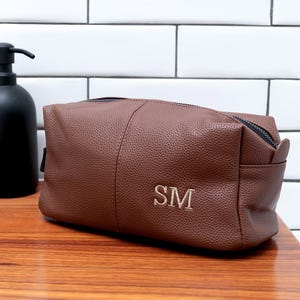 May include: Brown leather toiletry bag with the initials "SM" embroidered in gold. The bag has a zipper closure and a small side pocket. The bag is sitting on a wooden surface.