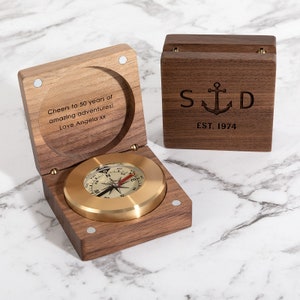 May include: A wooden compass in a hinged box, with a brass compass face. The box is engraved with "Cheers to 50 years of amazing adventures! Love Angela xx." The lid of the box is open, revealing the compass. A separate wooden box is also present, engraved with "S D" and an anchor, and "EST. 1974."