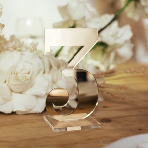May include: A gold-coloured number 5 table decoration with a clear acrylic base. The number has a reflective surface and is displayed in front of white flowers and a wooden surface. Suitable for weddings or special events.