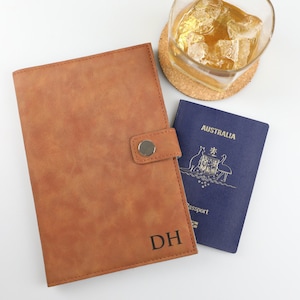 May include: A tan leather-effect passport holder featuring a snap button closure and the embossed initials DH in the bottom right corner, positioned next to a blue Australian passport and a glass of amber liquid on a round cork coaster.