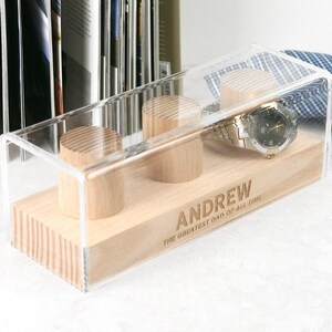 May include: A clear acrylic watch box with a wooden base. The box has three compartments for watches and a watch is displayed in one of the compartments. The base is engraved with the name "ANDREW" and the text "THE GREATEST DAD OF ALL TIME".