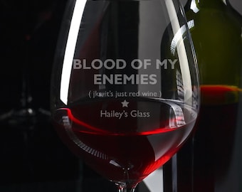 May include: A clear wine glass filled with red wine, next to a green wine bottle. The glass has the text "BLOOD OF MY ENEMIES (jk it's just red wine)" and "Hailey's Glass" etched on it. The glass is on a reflective black surface.