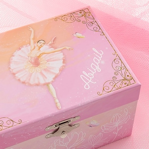 May include: Pink ballerina music box with a ballerina illustration and the name "Abigail". The box features a hinged lid, a silver clasp, and gold decorative accents. The background has a gradient of pink and peach colors.