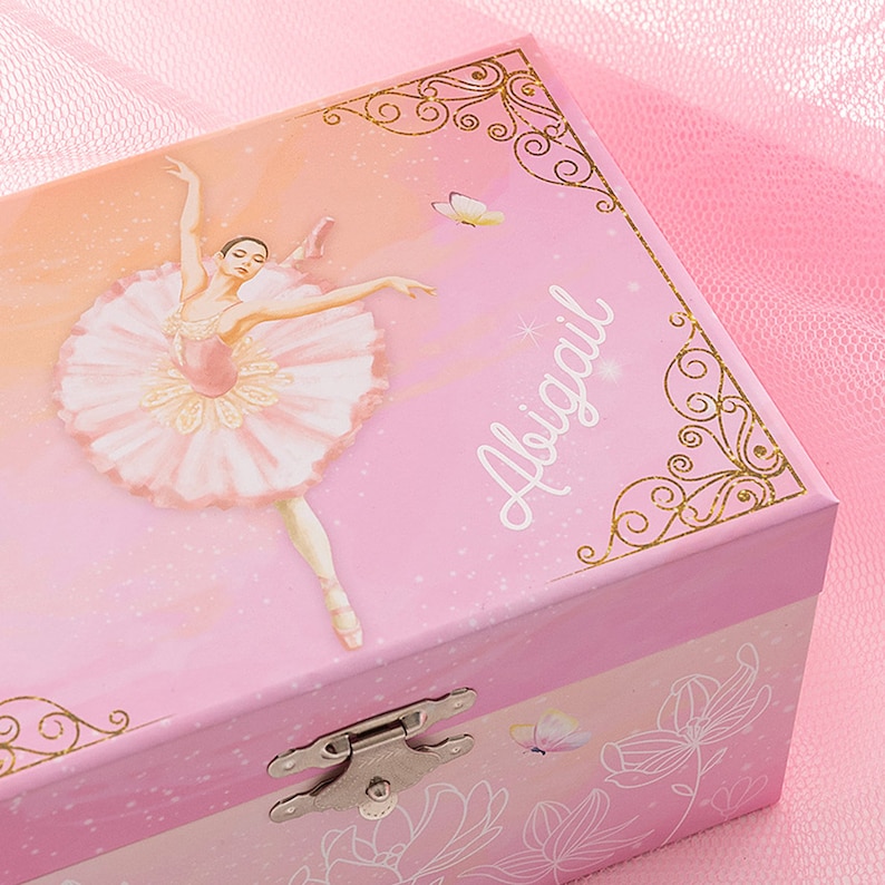 May include: Pink ballerina music box with a ballerina illustration and the name "Abigail". The box features a hinged lid, a silver clasp, and gold decorative accents. The background has a gradient of pink and peach colors.