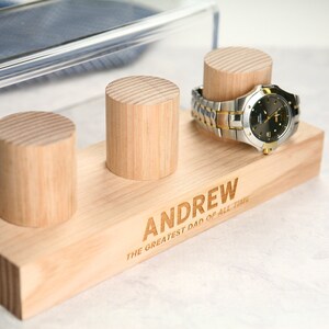 May include: Wooden watch stand with three slots for watches. The stand is engraved with the text "ANDREW THE GREATEST DAD OF ALL TIME".