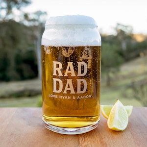 May include: A clear glass beer mug with a frosted rim and a white head of foam. The mug is filled with a golden-coloured beer and has the words "RAD DAD" etched on the side, along with the text "LOVE RYAN & AARON". The mug is sitting on a wooden surface with lemon slices beside it.