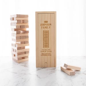 May include: A wooden tumbling tower game set with a tower of blocks and a wooden box. The box is engraved with "THE JOHNSON FAMILY" and "TUMBLING TOWER GAME". The game includes wooden blocks for building and playing.