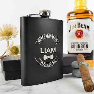 May include: Black stainless steel hip flask with a silver engraved design. The design includes the words "Groomsman Liam" with a bow tie and the date "11.03.2023".