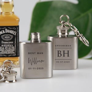 May include: Two silver-coloured hip flasks with keychains. One is engraved with "Best Man", a name, and a date. The other has "Groomsman", initials, and a date. A whiskey bottle is visible in the background.