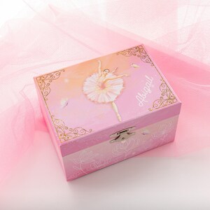 May include: A pink music box with a ballerina illustration and the name "Abigail". The box features a hinged lid, a silver clasp, and a floral design on the bottom. Gold filigree frames the ballerina image.