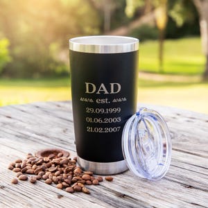 May include: A matte black stainless steel tumbler with a silver rim and base. The front is laser-engraved with the text DAD, est., and three dates: 29.09.1999, 01.06.2003, and 21.02.2007. A clear plastic lid sits beside the tumbler on the surface.