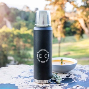 May include: A black insulated flask with a silver lid and base. The flask features a white circular logo with the letters "E" and "C". A bowl of soup and greenery are in the background.