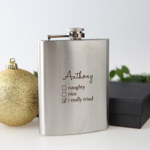 May include: A silver-coloured hip flask with a hinged cap. The flask is engraved with the name "Anthony" and a checklist that reads "naughty", "nice", and "i really tried". A gold Christmas bauble and a black gift box are in the background.