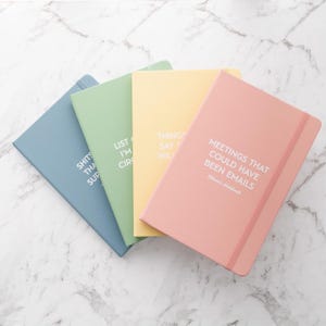 May include: Four notebooks in pastel shades of blue, green, yellow, and pink, displayed on a marble surface. Each notebook features white text on the cover, with phrases like "LIST I'M CIR" and "MEETINGS THAT COULD HAVE BEEN EMAILS".