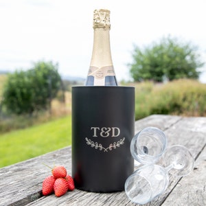 May include: A black insulated cooler with a personalised engraving of "T&D" and a leafy design. The cooler is holding a bottle of champagne and is sitting on a wooden table with two wine glasses and strawberries.