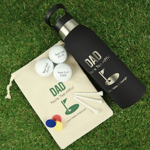 May include: A black water bottle with a silver cap and the text DAD You're Tee-riffic! with a golf hole graphic and Love Helen & Georgia. Includes a beige drawstring bag with matching text, three white golf balls, three white tees, and three markers.