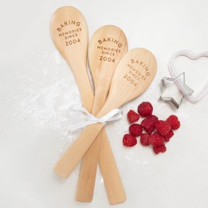 May include: Three wooden spoons with the text "Baking Memories Since 2004" engraved on them. The spoons are tied together with a white ribbon and are surrounded by red raspberries and white powder.