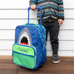 May include: Blue rolling backpack with a shark design. The backpack features a large shark face on the front, a green front pocket with the name "Oakley", and a blue handle. The bag has a shark pattern.