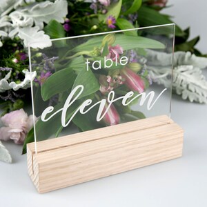 May include: Clear acrylic table number sign with the text "table eleven" in white script font. The sign is on a wooden stand.