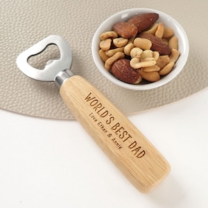 May include: A bottle opener with a light-coloured wooden handle featuring the engraved text "WORLD'S BEST DAD" and "Love Ethan & Annie". The metal opener head is silver-toned and polished, positioned next to a small white bowl filled with mixed nuts.