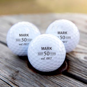 May include: Three white golf balls, each printed with "MARK", "AGED 50 YEARS", and "est. 1977" in black. The golf balls are resting on a weathered wooden surface.