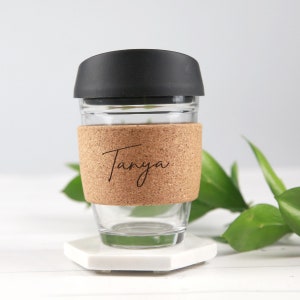 May include: A clear glass travel mug with a black lid and a cork sleeve. The name "Tanya" is printed on the cork sleeve in a handwritten style font. The mug is sitting on a white hexagonal coaster.