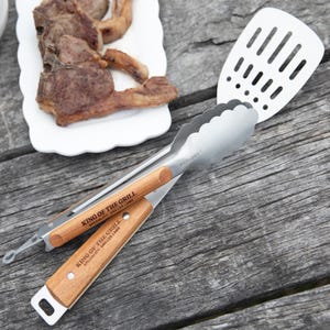 May include: Grilling tongs and a spatula with wooden handles, alongside grilled lamb chops on a white plate. The tongs are branded "KING OF THE GRILL" and "ECOLOGY". The scene suggests outdoor cooking.