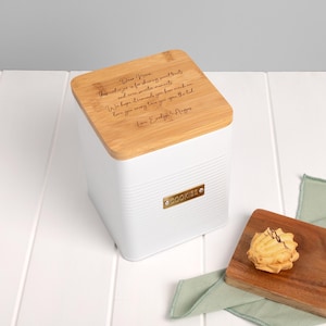 May include: White cookie jar with a bamboo lid. The lid has engraved text. A gold-colored label on the jar reads "COOKIES". A cookie sits on a wooden cutting board next to the jar. The jar is a square shape.