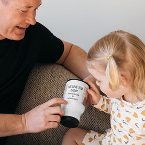 May include: A white travel mug with a black lid and base, featuring a printed design that reads WE LOVE YOU DADDY, HAPPY FATHER'S DAY, and JAZMIN & CHARLI, along with a small heart icon and two vertical photo panels on the sides of the text.