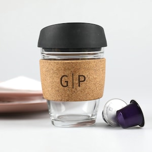 May include: A clear glass travel mug with a black lid and a cork sleeve. The cork sleeve has the initials "G|P" etched into it. A purple and silver coffee pod rests next to the mug.