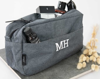 May include: Grey zipped wash bag with a white monogram "MH" embroidered on the front. The bag is filled with various toiletries, including a bottle of cologne, a bottle of body wash, and a small bottle of essential oil.