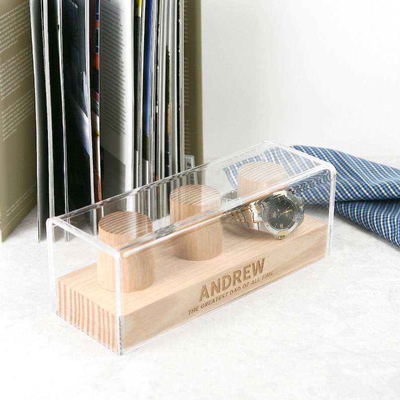 May include: A clear acrylic watch box with a wooden base. The box has three compartments for watches and a watch is displayed in one of the compartments. The base is engraved with the name "ANDREW" and the text "THE GREATEST DAD OF ALL TIME".