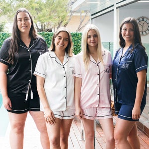 May include: Four people wearing matching pajama sets. The sets include a short-sleeved button-down shirt and shorts, all trimmed with white piping. The pajamas are in black, white, pink, and navy blue. Each shirt has embroidered initials.