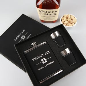 May include: A black hip flask and shot glass set with the text "THIRST AID" and "MIKE ANDREWS" in white. The set includes a flask and two shot glasses, presented in a black gift box. A bottle of bourbon and a bowl of pistachios are in the background.