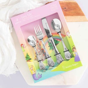 May include: A four-piece children's cutlery set in a box with a fairy tale theme. The set includes a knife, fork, spoon, and teaspoon. The box has illustrations of a castle, mermaid, and fairy. The cutlery is stainless steel.