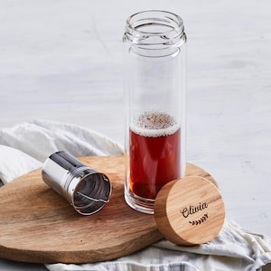 May include: A clear glass water bottle with a stainless steel infuser and a wooden lid engraved with the name "Olivia" and a leaf design. The bottle contains a reddish-brown liquid. The items are on a wooden cutting board.