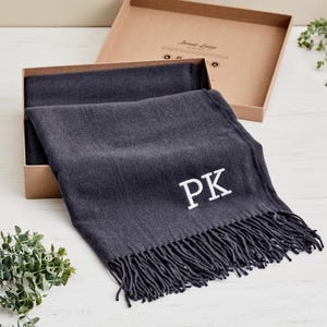 May include: A charcoal grey scarf with white embroidered initials "PK" and a fringed edge, presented in a brown gift box. The scarf is partially visible, suggesting a gift. The box has text on the lid.