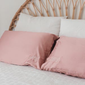 May include: Two blush pink pillowcases with embroidered initials, placed on a white bed with a textured cover. The pillows are in front of two white pillows and a woven headboard.