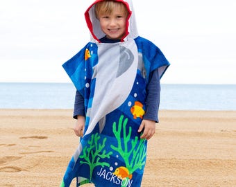May include: A blue shark-themed hooded beach towel with a white shark face hood with red trim. The towel features an underwater scene with fish and coral. The name "JACKSON" is printed on the towel.
