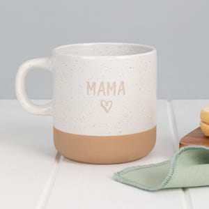 May include: White ceramic mug with a speckled finish and a tan base. The word "MAMA" is printed in light brown, with a heart symbol below. The mug has a curved handle and is set on a white surface.